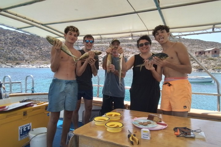 Five people on a boat holding fish, with a sea and mountain background.