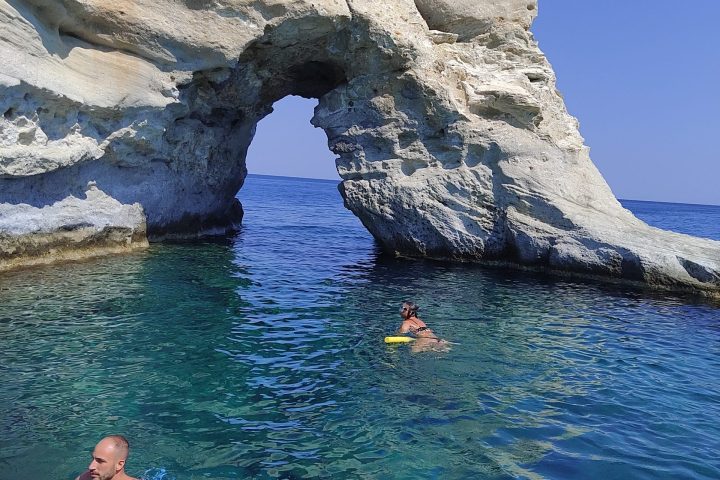 Two people swimming near a natural rock arch in clear blue sea.