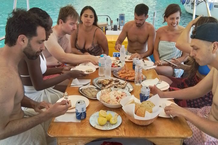 Group of people on a boat enjoying a meal with seafood by turquoise water.